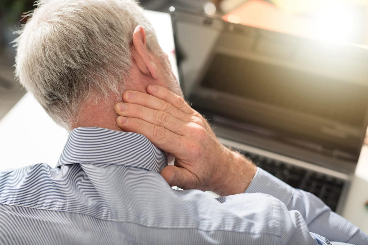 Older man looking down at a computer has his hand on that back of his neck that is in pain.
